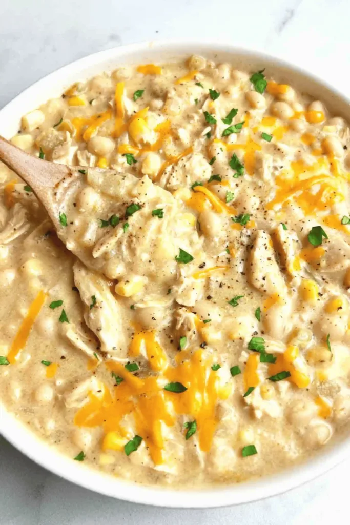 Portrait bowl of white chicken chili topped with avocado, cilantro, and tortilla strips beside lime wedges and cornbread